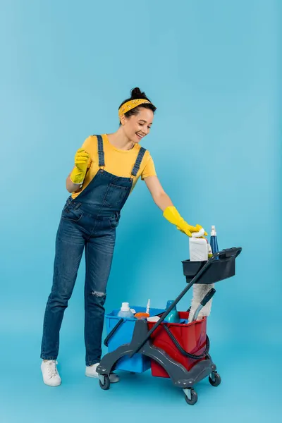 Jovem limpador em macacão jeans sorrindo perto do carrinho com suprimentos de limpeza em azul — Fotografia de Stock