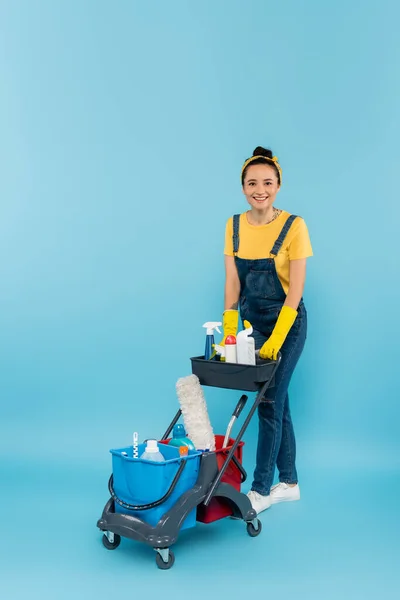 Happy cleaner in denim overalls looking at camera near trolley with cleaning supplies on blue — Stock Photo