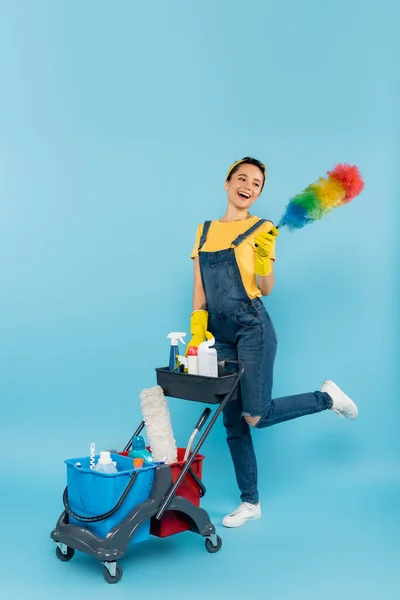 Cheerful cleaner with dust brush posing near cart with detergents and buckets on blue — Stock Photo