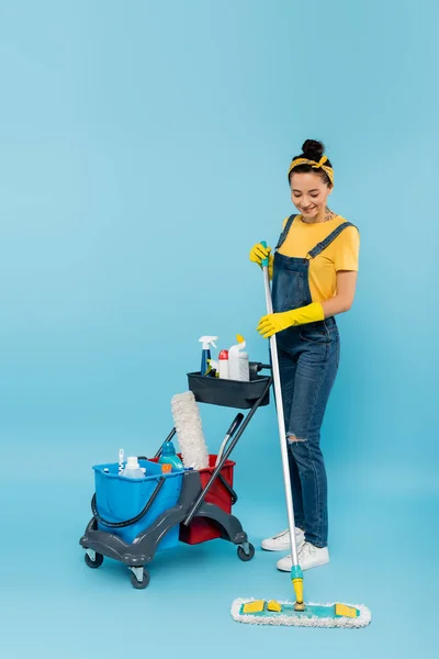 Nettoyant souriant avec balai près du chariot avec seaux et détergents sur bleu — Photo de stock