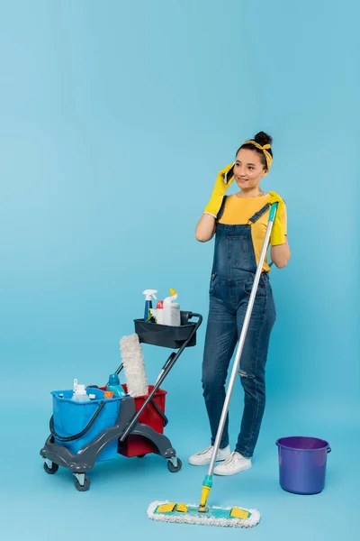 Reiniger mit Wischmopp spricht auf Smartphone in Warenkorb mit Reinigungsmitteln auf blau — Stockfoto