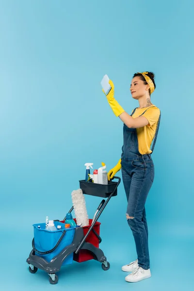 Detergente in tuta denim e guanti di gomma scattando selfie vicino al carrello con prodotti per la pulizia su blu — Foto stock