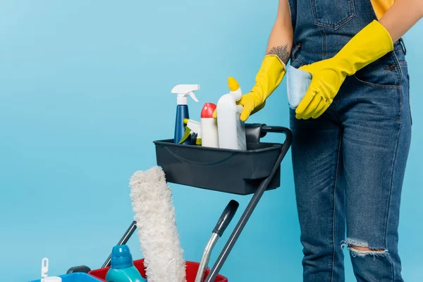 Vista cortada de limpador em luvas de borracha segurando esponja perto do carrinho com suprimentos de limpeza isolados em azul — Fotografia de Stock