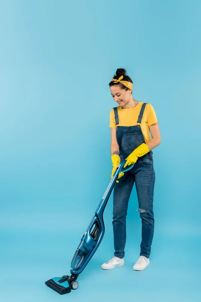 Sorridente dona de casa em macacão jeans e luvas de borracha amarela aspirando em azul — Fotografia de Stock