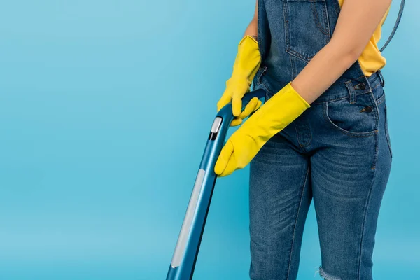 Vista parziale di casalinga in tuta denim e guanti di gomma gialla aspirapolvere isolato su blu — Foto stock