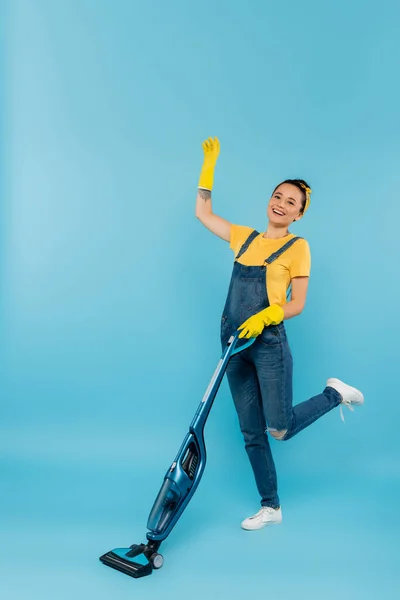 Cheerful woman in denim clothes and rubber gloves waving hand while vacuuming on blue — Stock Photo