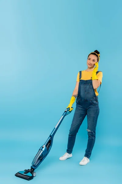 Woman in denim overalls and rubber gloves talking on smartphone while vacuuming on blue — Stock Photo