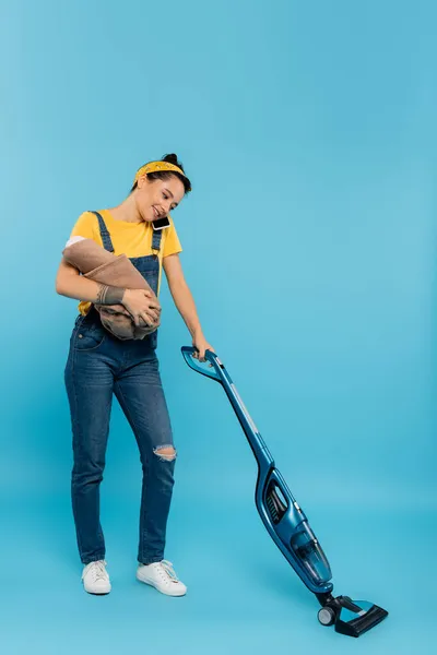 Happy woman with baby doll talking on mobile phone while vacuum uming on blue — стоковое фото