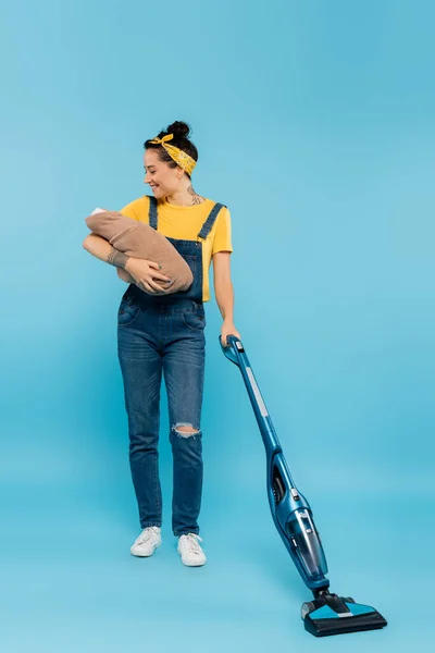 Joyful woman in denim overalls holding baby doll while vacuuming on blue — Stock Photo