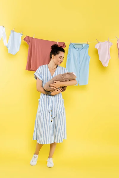 Felice casalinga tenendo bambola bambino vicino lavanderia appeso sulla corda su sfondo giallo — Foto stock