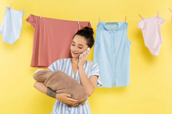 Mujer sonriente sosteniendo la muñeca mientras habla en el teléfono inteligente cerca de colgar la ropa en amarillo - foto de stock