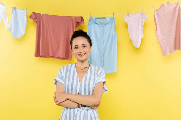 Happy housewife looking at camera while standing with crossed arms near hanging laundry on yellow background — Stock Photo