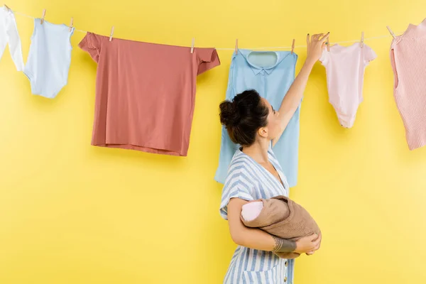 Brunette woman hanging clothing on rope while holding baby doll on yellow background — Stock Photo