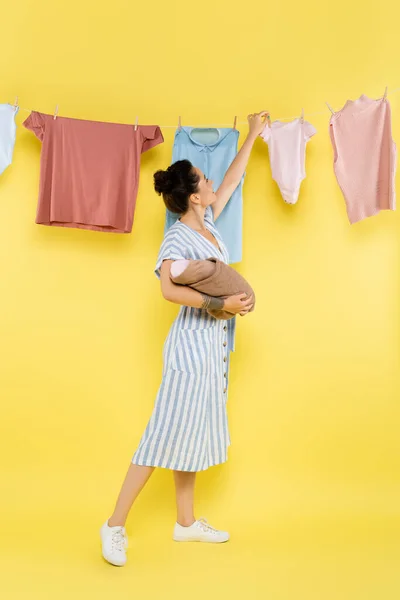 Jovem dona de casa com bebê boneca pendurado lavanderia na corda no fundo amarelo — Fotografia de Stock