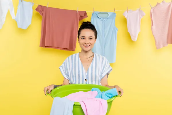 Joyful housewife holding laundry bowl near clothes hanging on rope on yellow background — Stock Photo