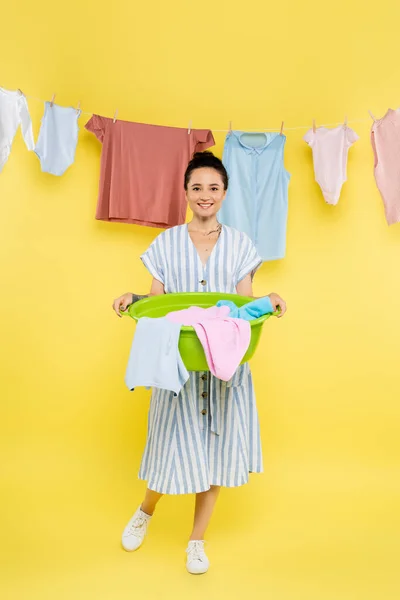 Full length view of smiling housewife holding laundry bowl near clothes hanging on yellow background — Stock Photo