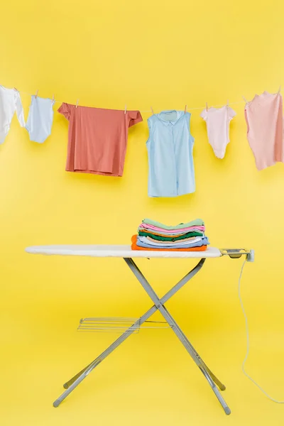 Pila de ropa limpia en tabla de planchar debajo de la ropa colgada en la cuerda sobre fondo amarillo - foto de stock