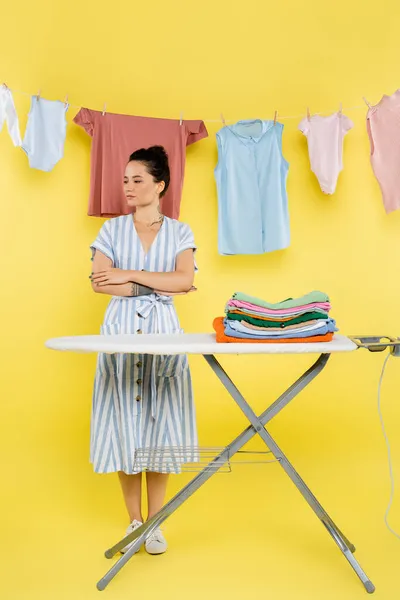 Brunette woman standing with crossed arms near stack of clothes on ironing board on yellow — Stock Photo