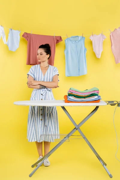 Brunette femme debout avec les bras croisés près de la pile de vêtements sur planche à repasser sur jaune — Photo de stock