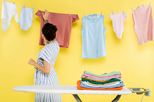 Morena mulher tocar pendurado lavanderia perto de tábua de engomar no amarelo — Fotografia de Stock
