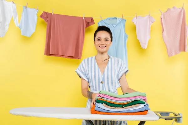 Fröhliche Hausfrau blickt in die Kamera neben Kleiderstapel auf Bügelbrett auf gelb — Stockfoto