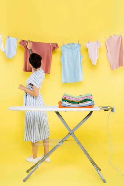 Brünette Frau steht neben hängender Kleidung und Bügelbrett auf gelb — Stockfoto