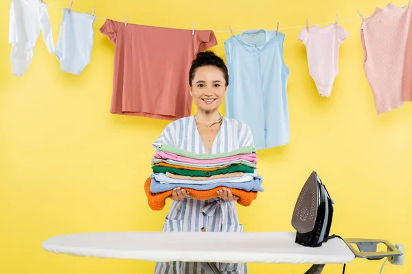 Mulher feliz olhando para a câmera enquanto segurando roupas perto de tábua de engomar no amarelo — Fotografia de Stock