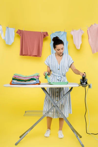 Brünette Frau hält Bügeleisen in der Nähe von Kleidung auf Bügelbrett auf gelb — Stockfoto
