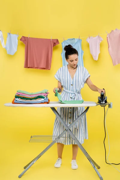 Morena mujer sosteniendo hierro cerca de la ropa en tabla de planchar en amarillo — Stock Photo