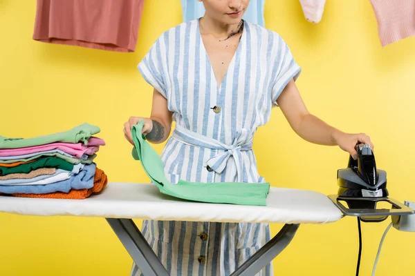 Частичный вид домохозяйки с одеждой и утюгом рядом с гладильной доской на желтом — стоковое фото