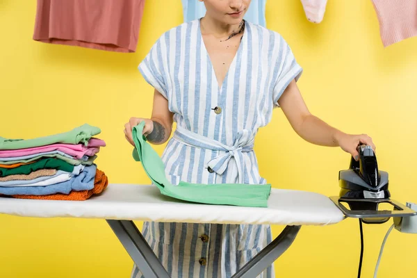 Vista parcial de ama de casa con ropa y plancha cerca de tabla de planchar en amarillo - foto de stock