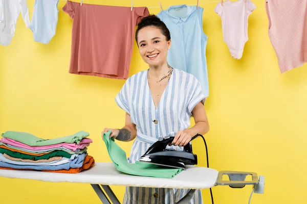 Brunetta donna sorridente a macchina fotografica mentre stirare vestiti vicino appeso lavanderia su giallo — Foto stock