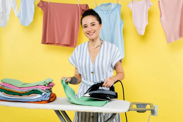 Mulher morena sorrindo para a câmera enquanto engomando roupas perto de pendurar roupa em amarelo — Fotografia de Stock