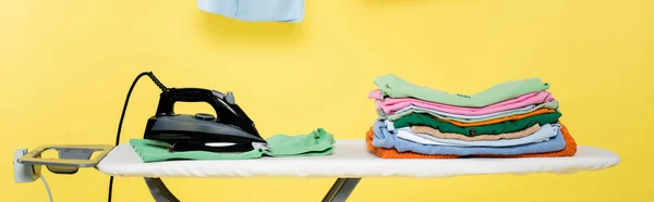 Tábua de engomar com pilha de lavandaria limpa e ferro elétrico no fundo amarelo, banner — Fotografia de Stock