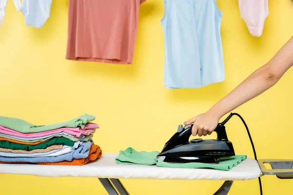 Vista recortada de ama de casa con plancha cerca de pila de ropa en tabla de planchar en amarillo - foto de stock