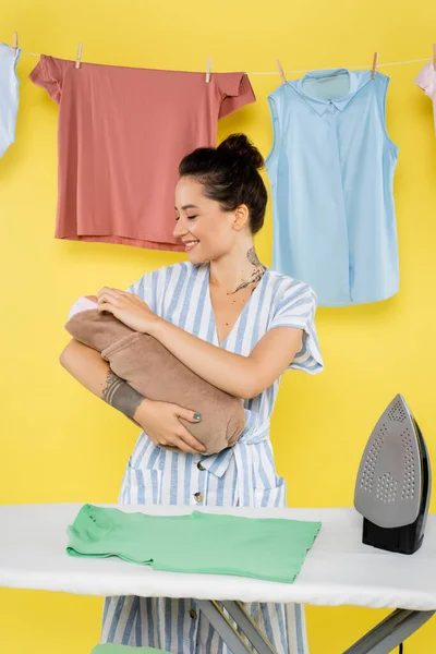 Cheerful woman holding baby doll near ironing board and laundry on yellow — Stock Photo