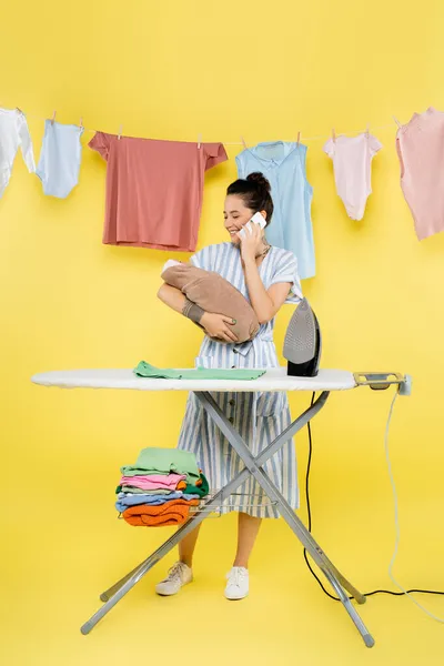 Glückliche Frau, die auf Smartphone spricht, während sie Babypuppe in der Nähe von Wäsche und Bügelbrett auf gelb hält — Stockfoto