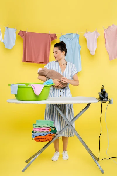 Brunetta donna tenendo bambino bambola vicino a lavanderia ciotola su asse da stiro su giallo — Foto stock