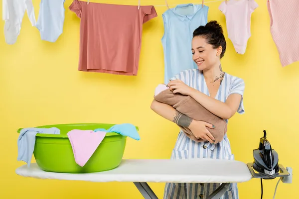 Sorrindo mulher segurando bebê boneca perto de lavandaria tigela em tábua de engomar no amarelo — Fotografia de Stock