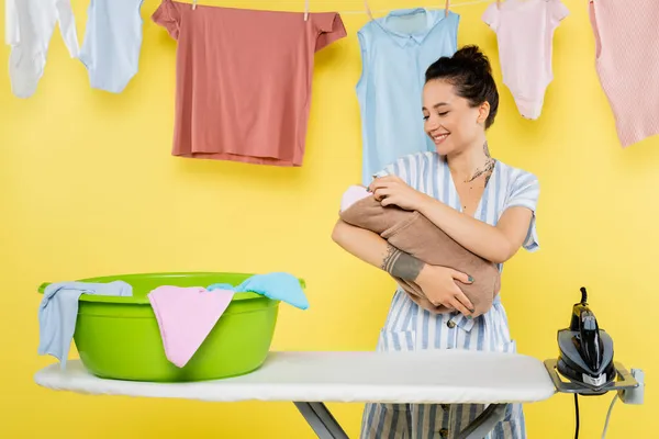 Donna sorridente che tiene la bambola vicino alla ciotola del bucato su asse da stiro su giallo — Foto stock