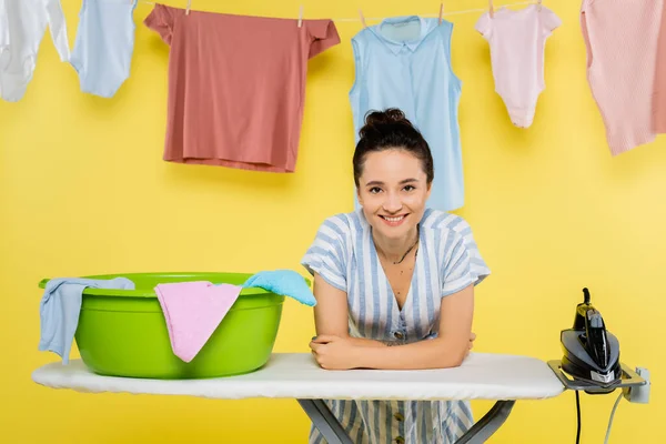 Happy housewife looking at camera while leaning on ironing board near laundry bowl on yellow — Stock Photo