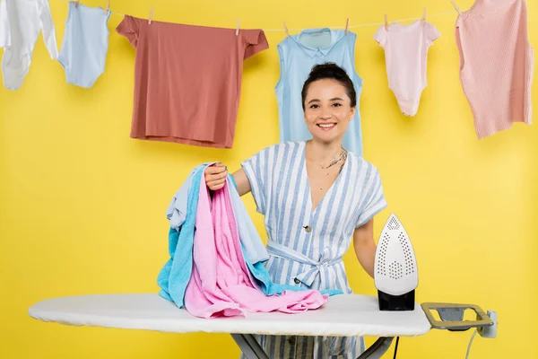 Femme au foyer souriante tenant buanderie près du fer à repasser sur planche à repasser sur jaune — Photo de stock