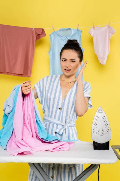 Müde Hausfrau blickt in die Kamera, während sie Kleidung in der Nähe von Bügelbrett auf gelb hält — Stockfoto