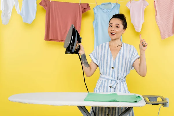 Happy housewife pointing at hanging laundry while holding iron near ironing board on yellow — Stock Photo