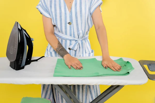 Vista recortada de mujer tatuada cerca de la ropa y plancha sobre tabla de planchar en amarillo - foto de stock