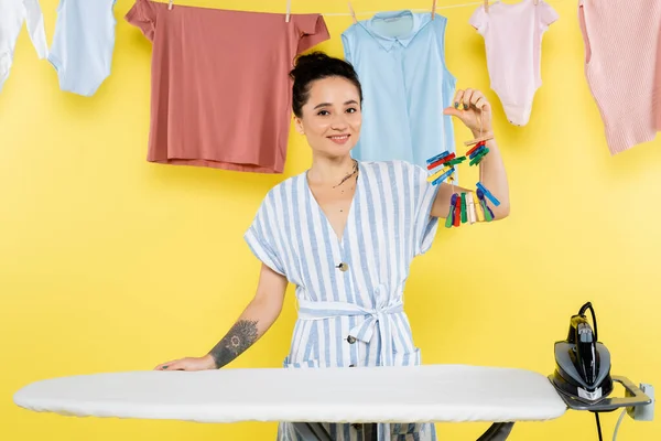 Татуированная женщина держит штифты для одежды возле гладильной доски и вешает белье на желтый — стоковое фото