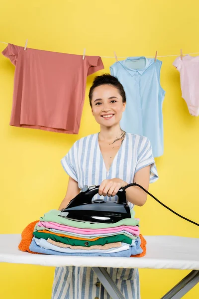 Pleased woman holding iron near stack of clothes on ironing board on yellow — Stock Photo