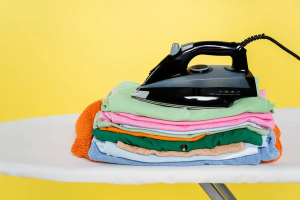 Stack of clean laundry and iron on ironing board isolated on yellow — Stock Photo
