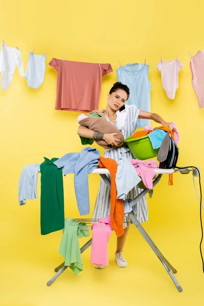 Woman talking on smartphone while holding baby doll and laundry bowl near pile of clothes on ironing board on yellow — Stock Photo