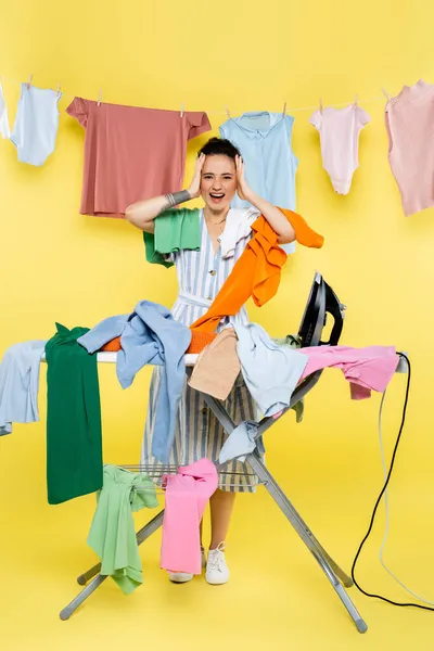 Shocked housewife touching head and screaming near clothing on ironing board on yellow — Stock Photo